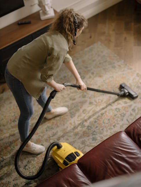 A woman with curly hair, dressed in a beige jacket and dark jeans, is performing surface cleaning with a vacuum cleaner on a patterned area rug in a living room. She is holding the vacuum's hose attac