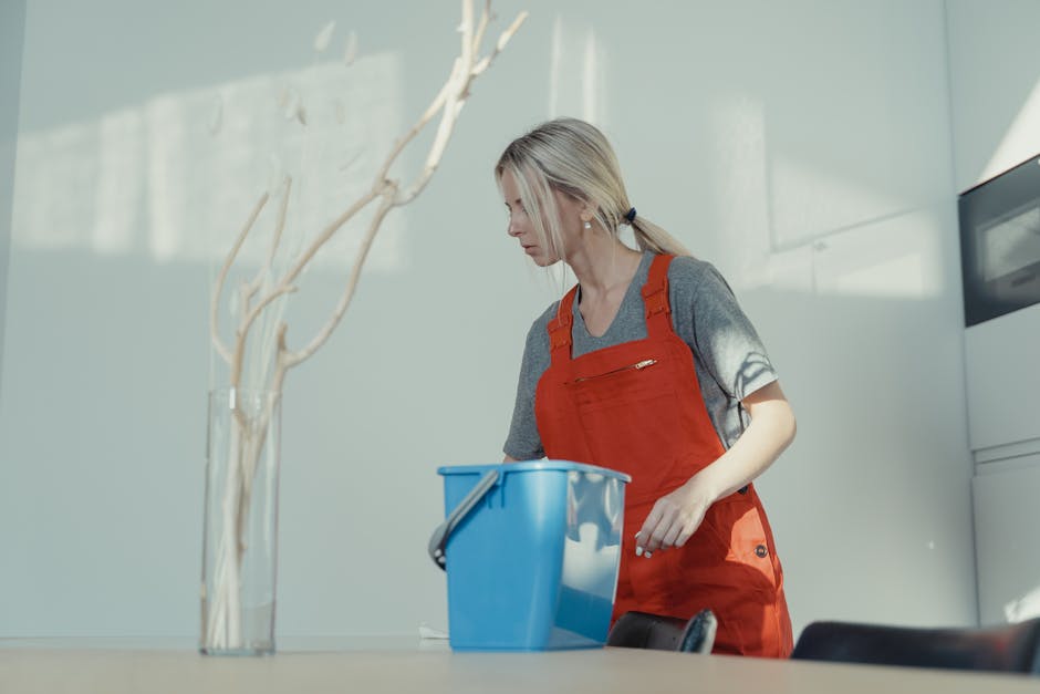 A woman wearing a grey t-shirt and an orange apron is engaged in surface cleaning in a well-lit indoor space. She has blonde hair tied back in a ponytail and appears focused on her task. The scene sho