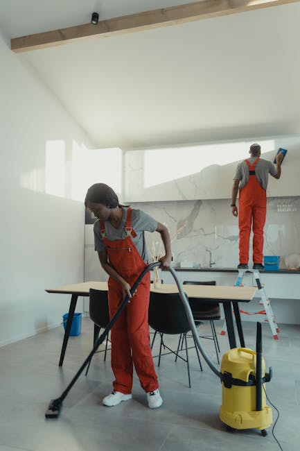 A woman wearing a grey t-shirt and an orange apron is engaged in surface cleaning in a well-lit indoor space. She has blonde hair tied back in a ponytail and appears focused on her task. The scene sho